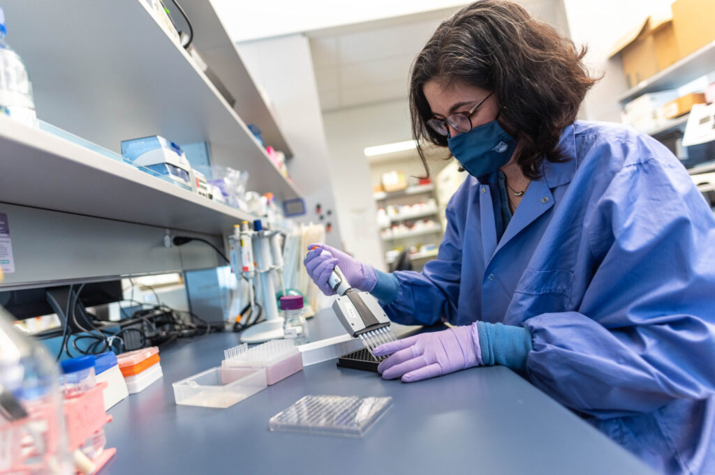 Publication author wearing a blue mask and lab coat sitting down and working in her lab.